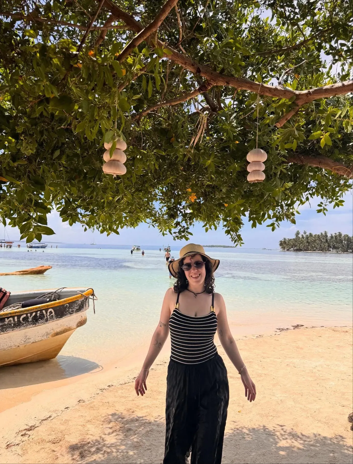 A traveler standing beneath a large tree on a beach in Guna Yala, with boats on the water behind