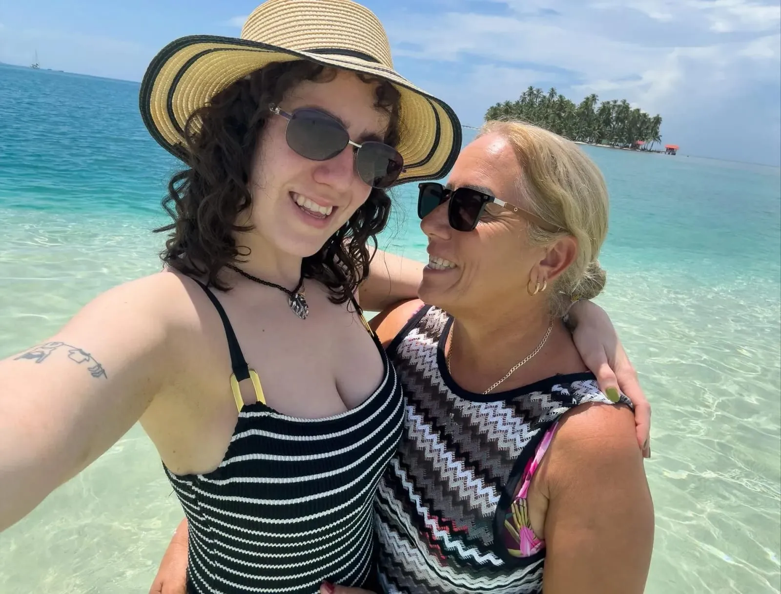 Two travelers smiling for a selfie in clear shallow water with a small palm island behind them