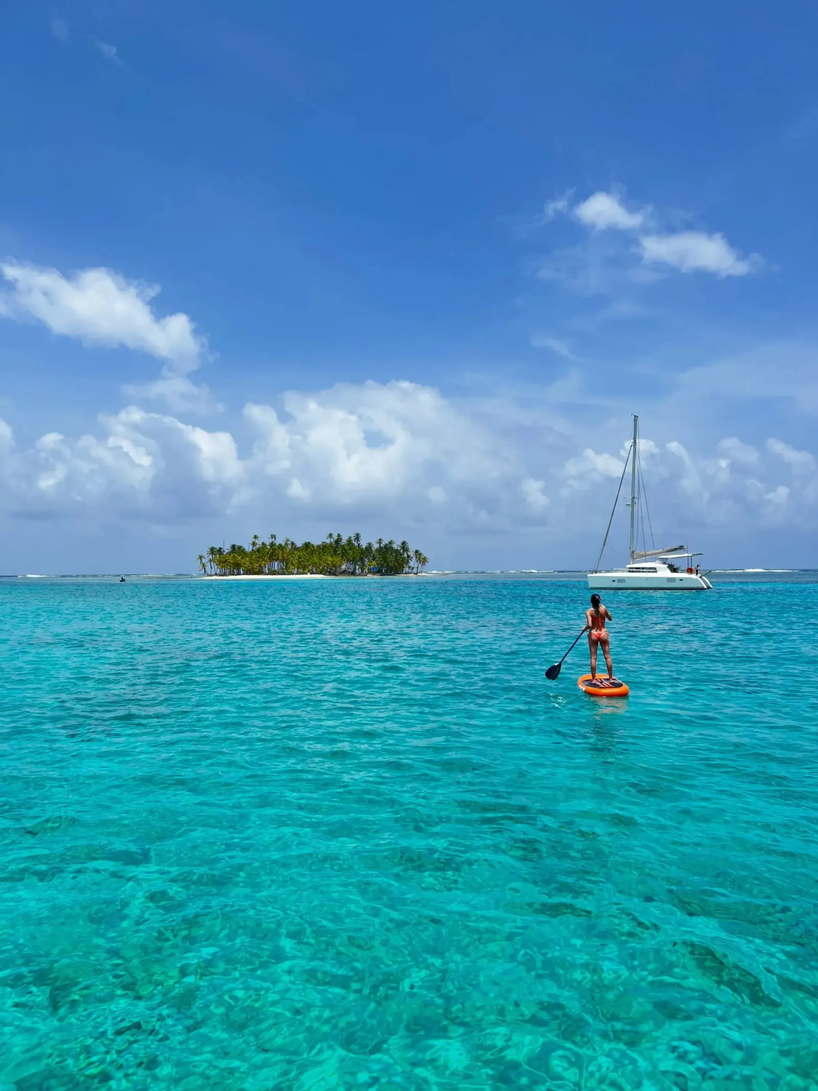 Una persona en tabla de paddle sobre agua turquesa, con una isla pequeña y un velero fondeado al fondo en Guna Yala